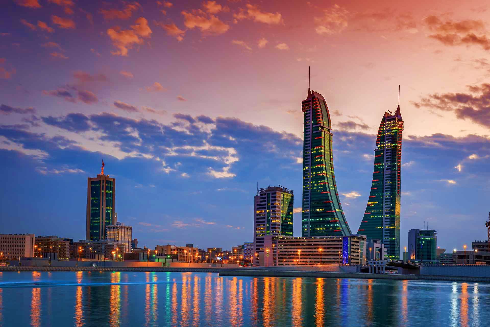 Bahrain Visa Bahrain skyline at sunset with illuminated skyscrapers reflected in blue water, featuring distinctive modern architecture tow